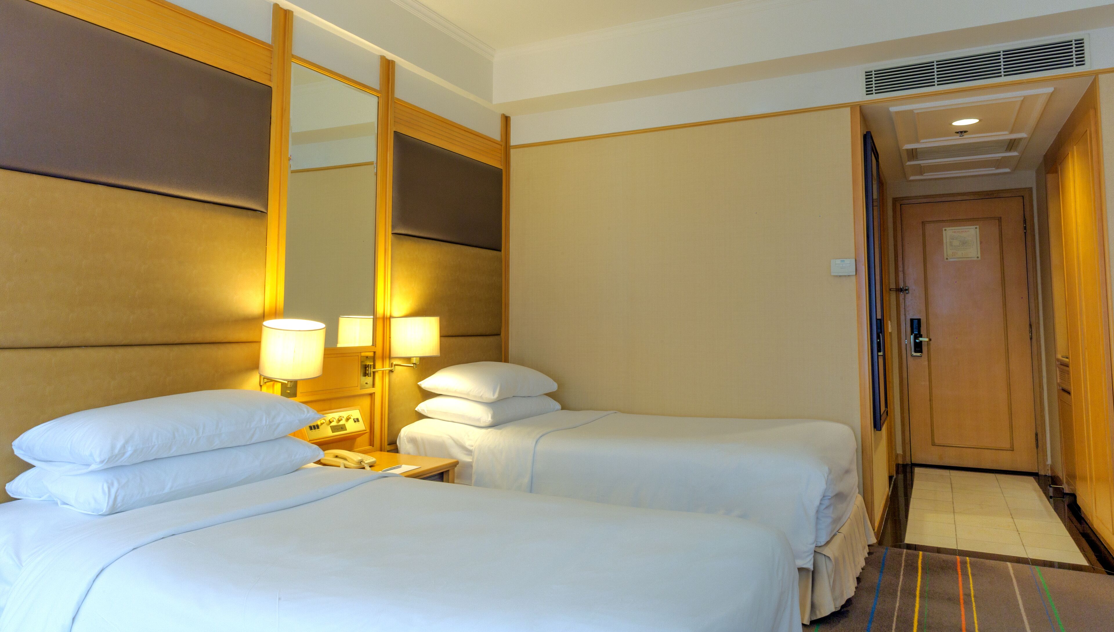 A hotel room with two beds, a bedside table with a lamp, a mirror, and a door leading to another room.