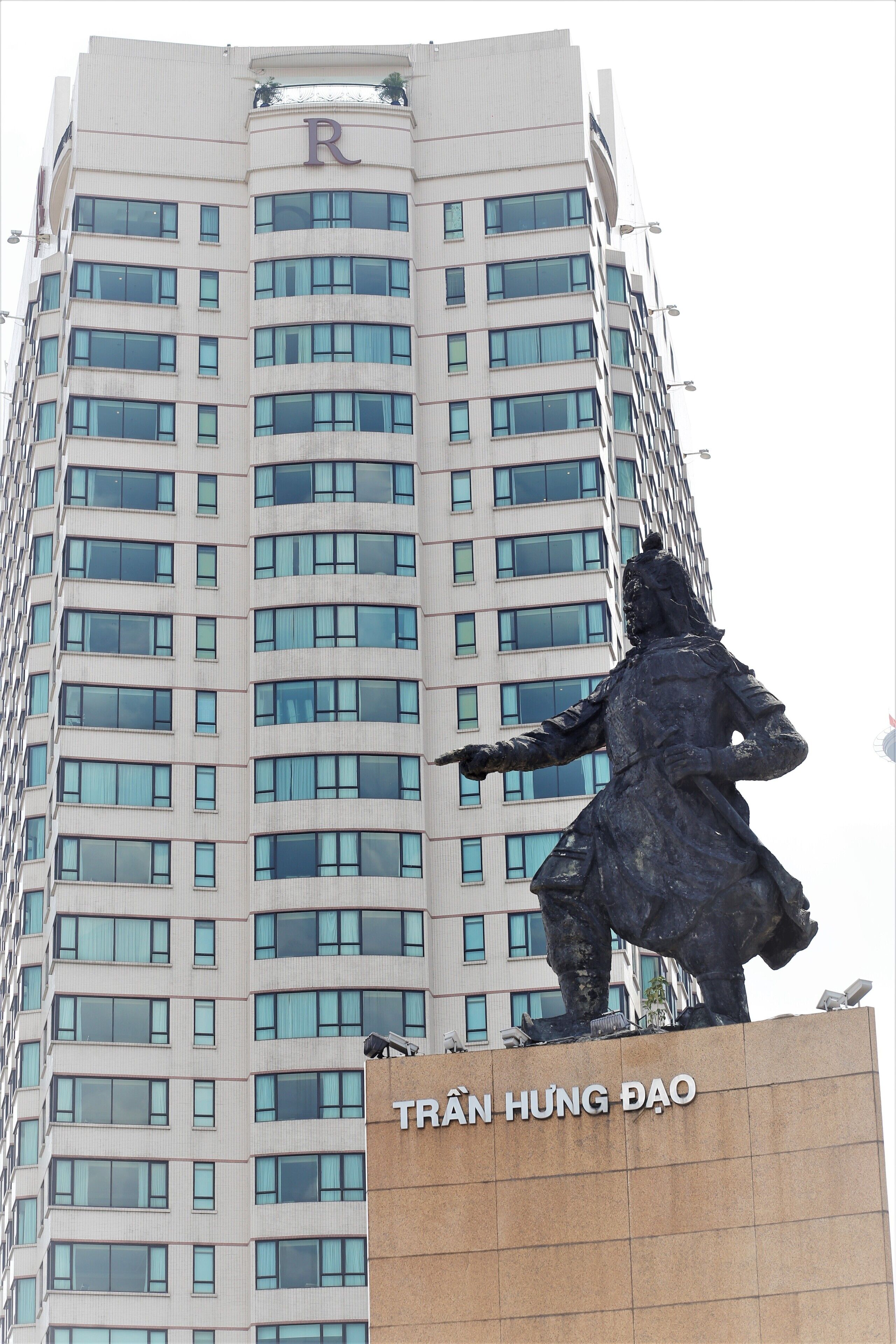 A tall building with a statue on top. The building has multiple windows and a sign that reads "TRÁN HUNG DAO".