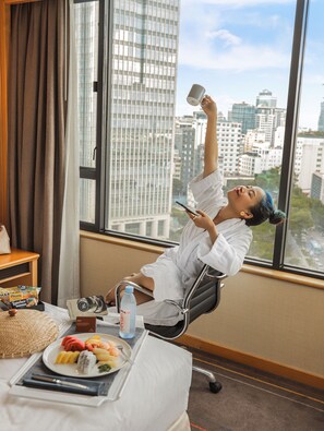 A person reclining in a chair by a window with a city view, a plate of food, and a bottle of water on a table. - Renaissance Riverside Hotel Saigon (Ho Chi Minh City)