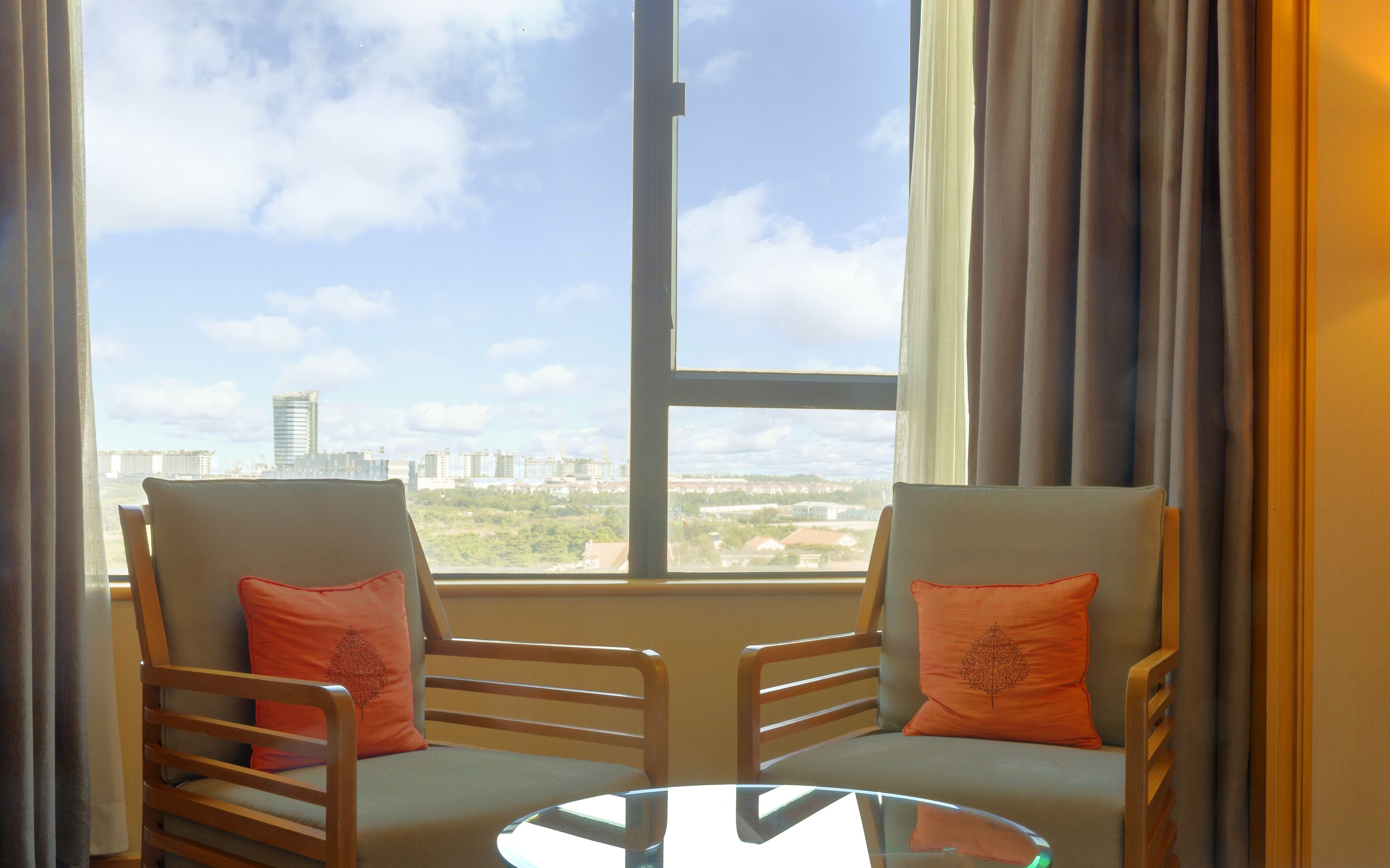 Two chairs with orange cushions, a glass table, and a large window with a view of buildings and trees.