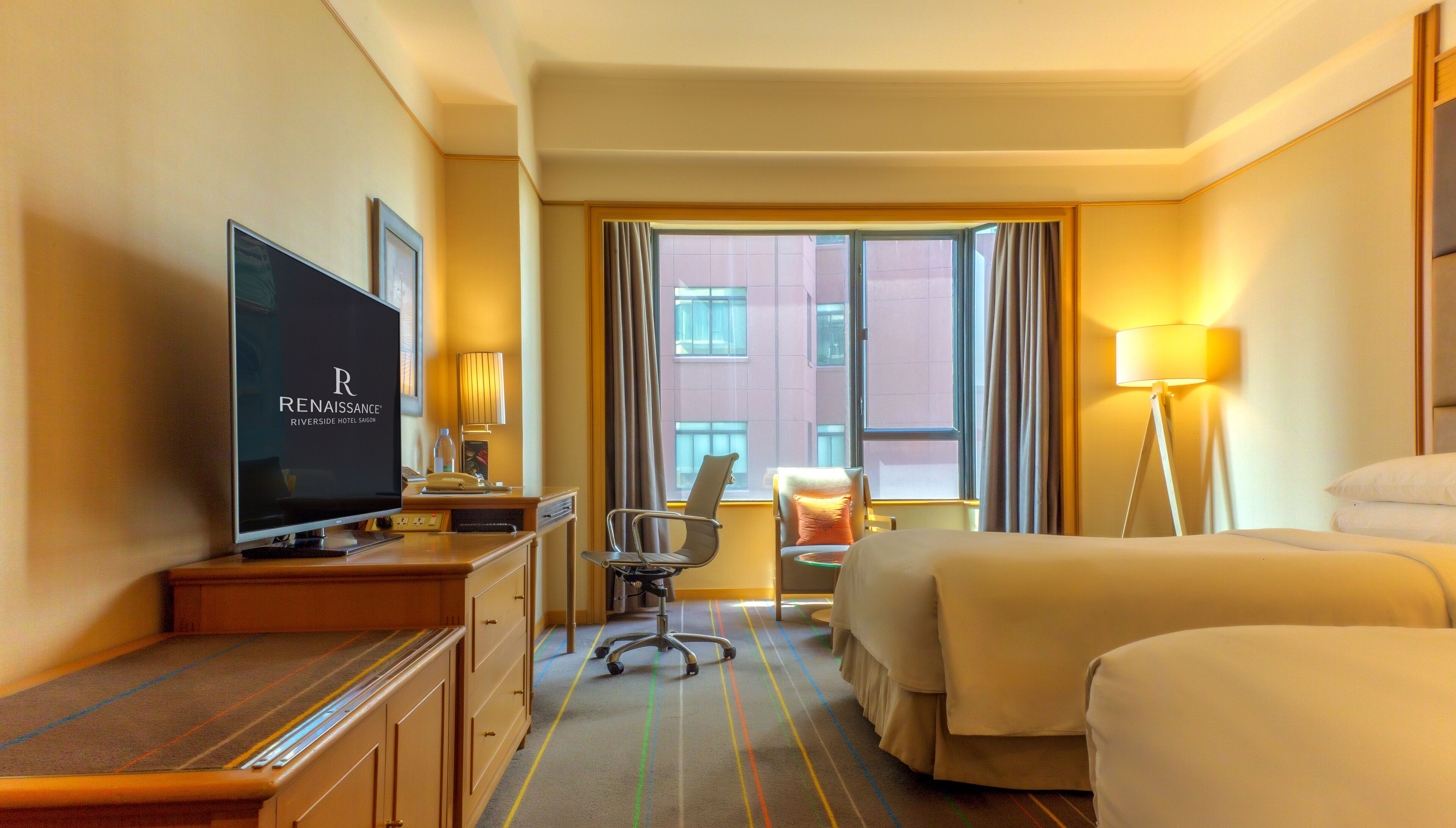 A hotel room with a television displaying the logo, a desk with a lamp, a chair, a bed, and a view of the city through the window.