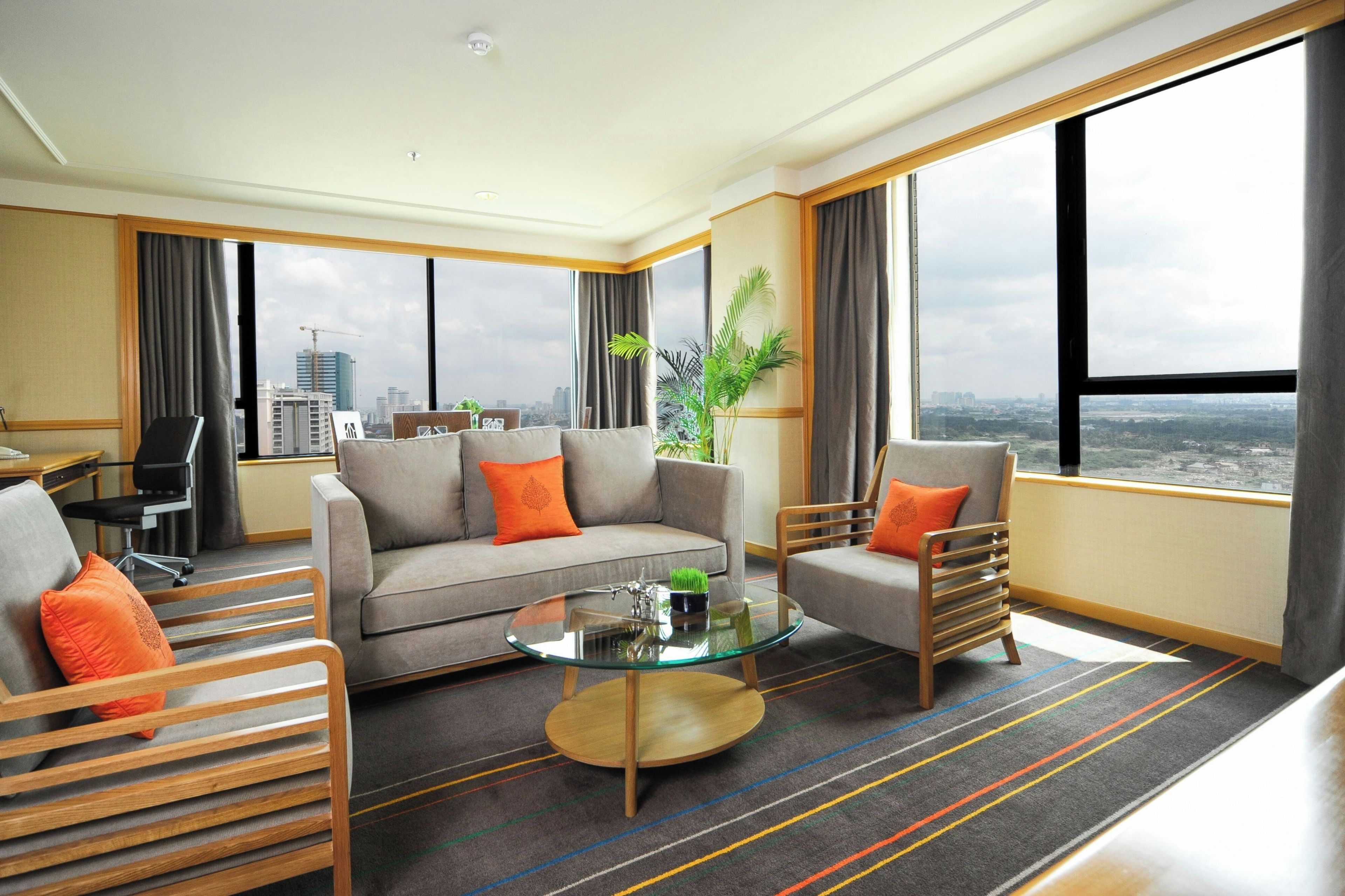 Une chambre d’hôtel moderne, dotée d’un canapé, de fauteuils, d’une table basse en verre et offrant une vue sur le paysage urbain.