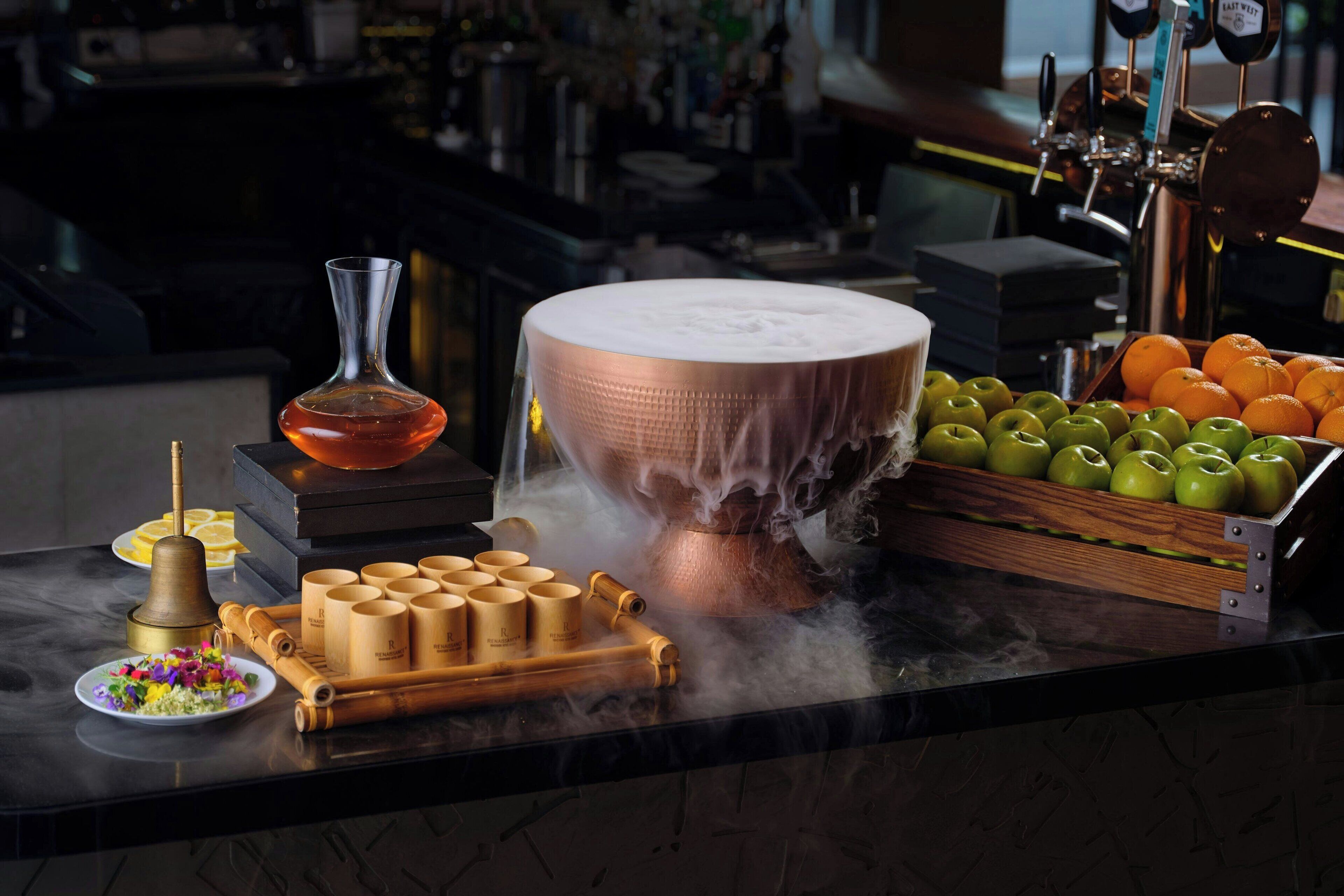 Una barra con una olla de cobre, un vaso, y una bandeja de madera con manzanas y naranjas.