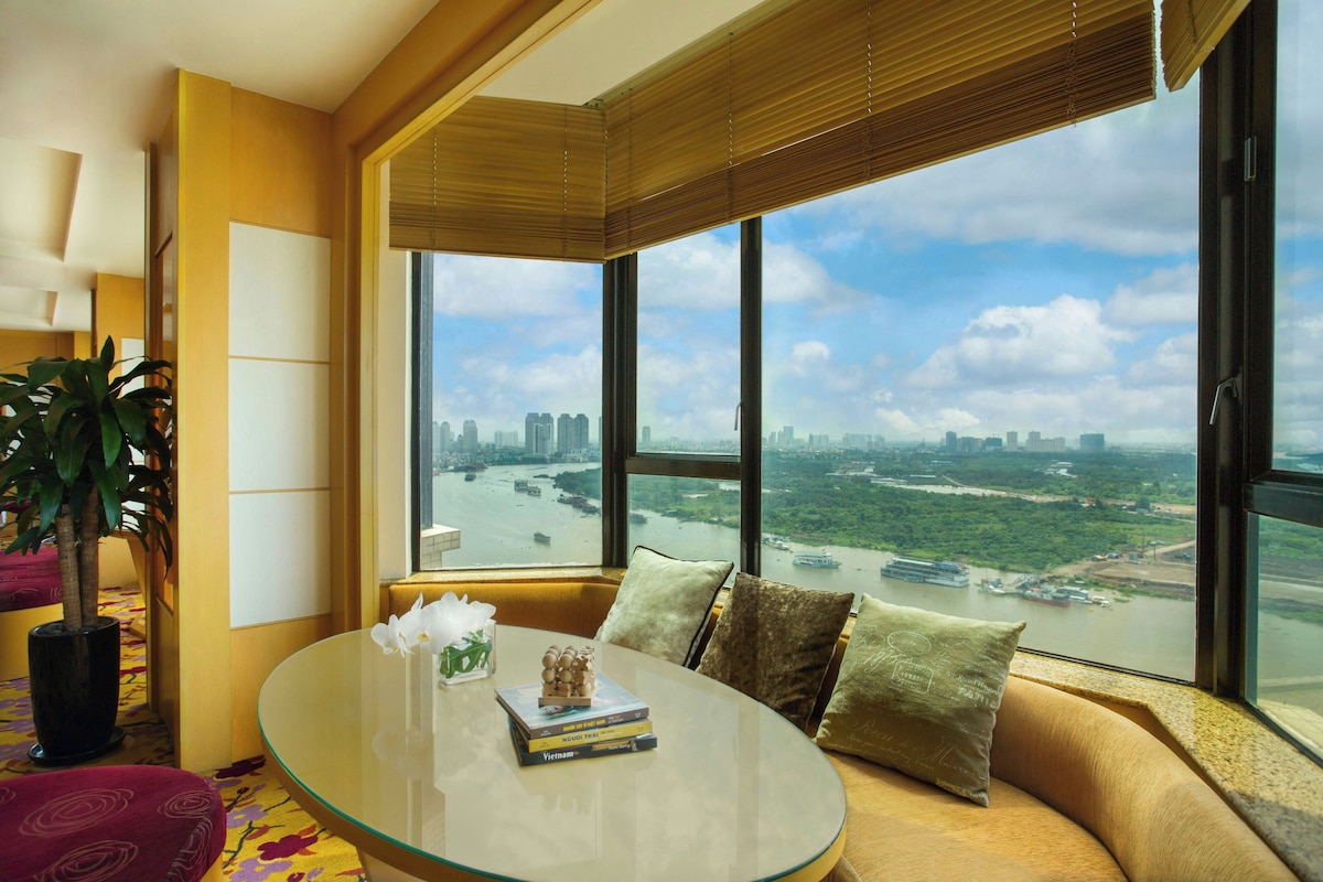 A modern living room with a glass coffee table, a sofa with cushions, a potted plant, and a large window offering a cityscape view.