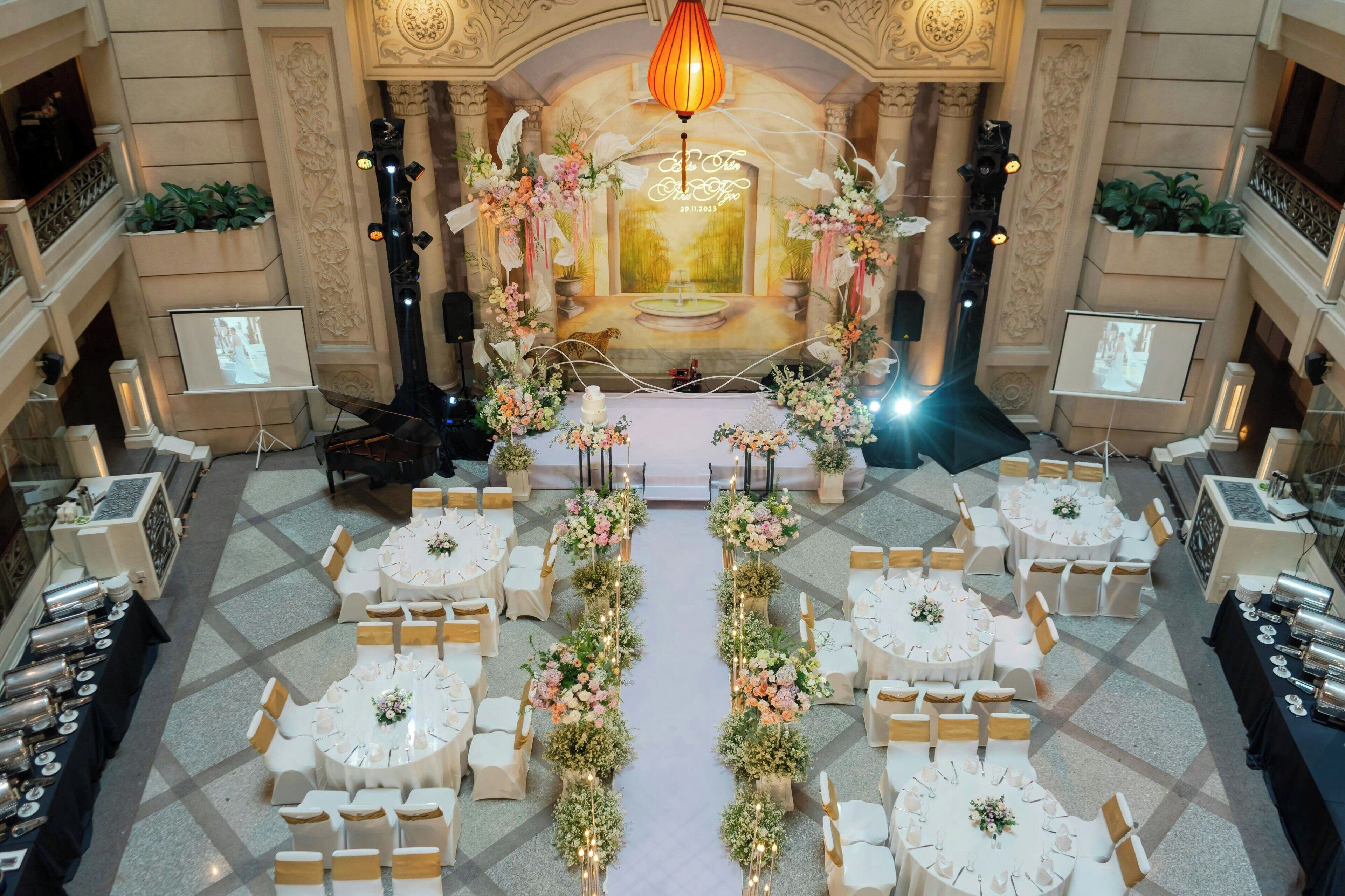 A grand ballroom with a central stage, surrounded by round tables with white tablecloths and gold chairs, and a floral aisle.