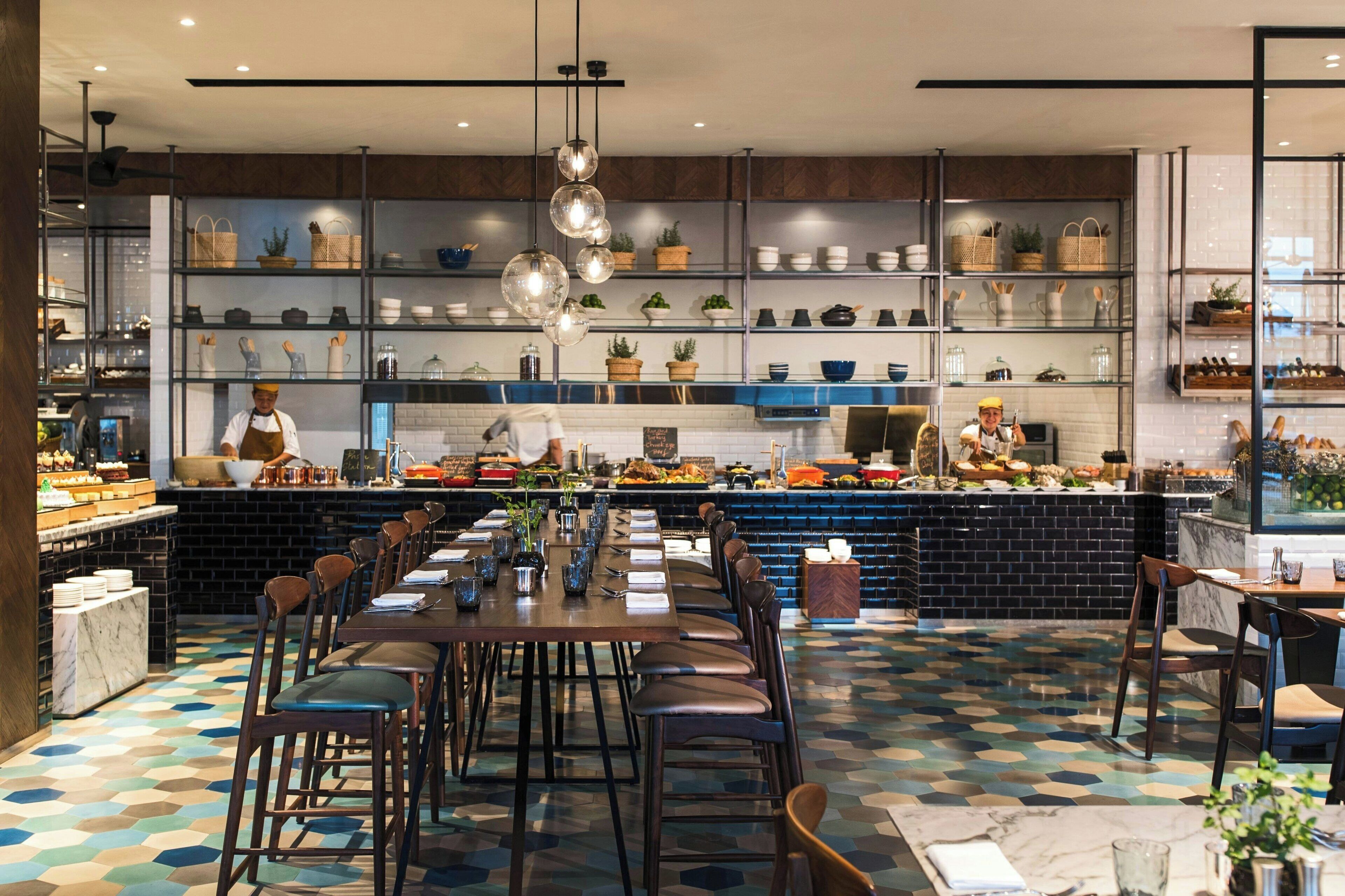 A restaurant with a long communal dining table, multiple seating options, and a kitchen area visible in the background.