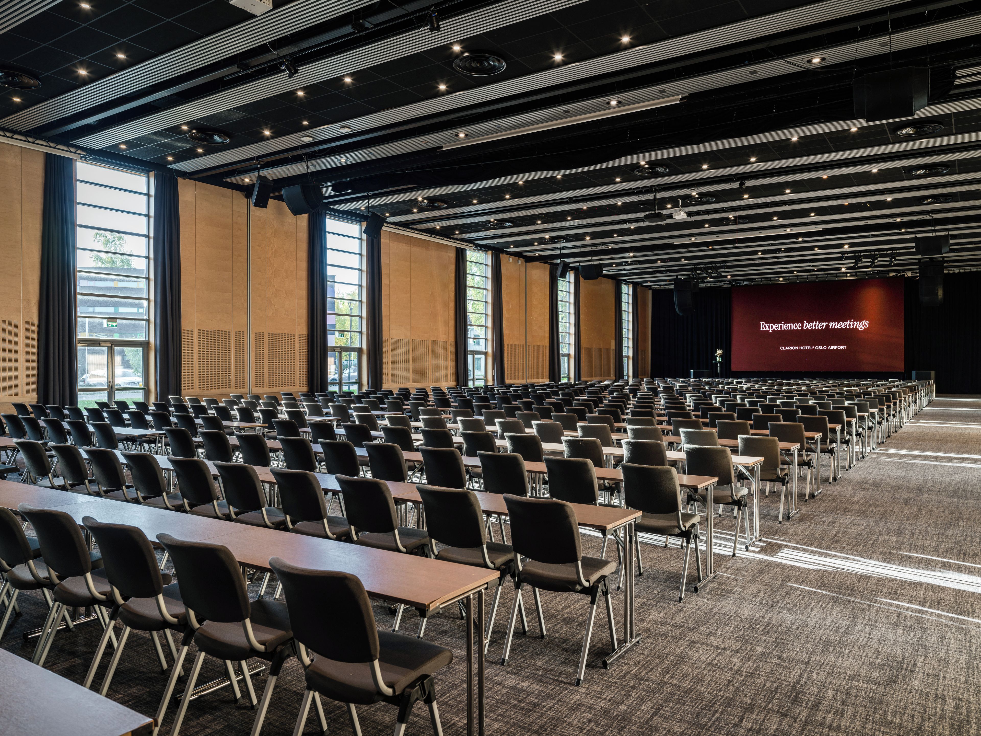 Ein Konferenzraum mit Reihen von Stühlen und Tischen, großen Fenstern und einer Bühne mit einem Bildschirm, der Text zeigt.