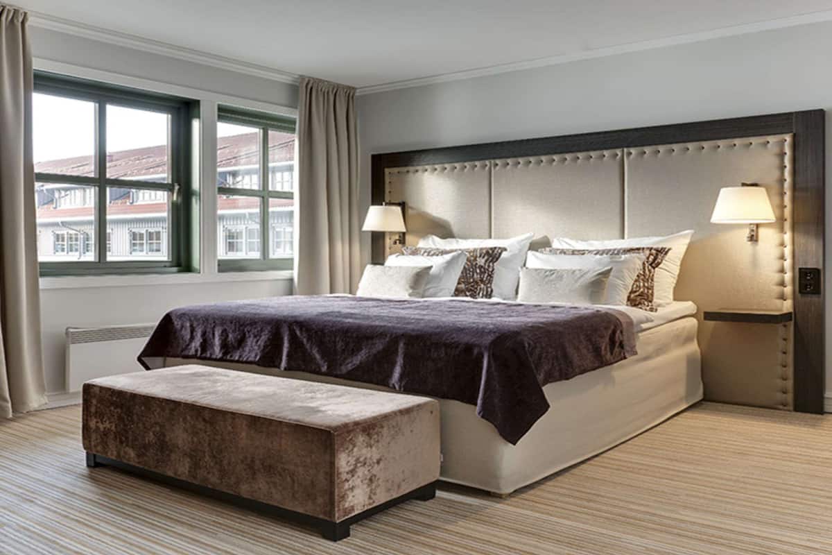 Habitación de hotel moderna con una cama grande, dos lámparas de noche, un banco y vistas a edificios a través de una ventana.