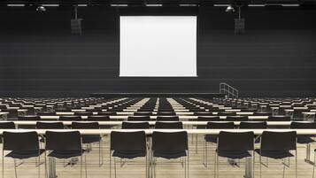 A large auditorium with rows of black chairs and tables, a large white screen at the front, and overhead lighting.