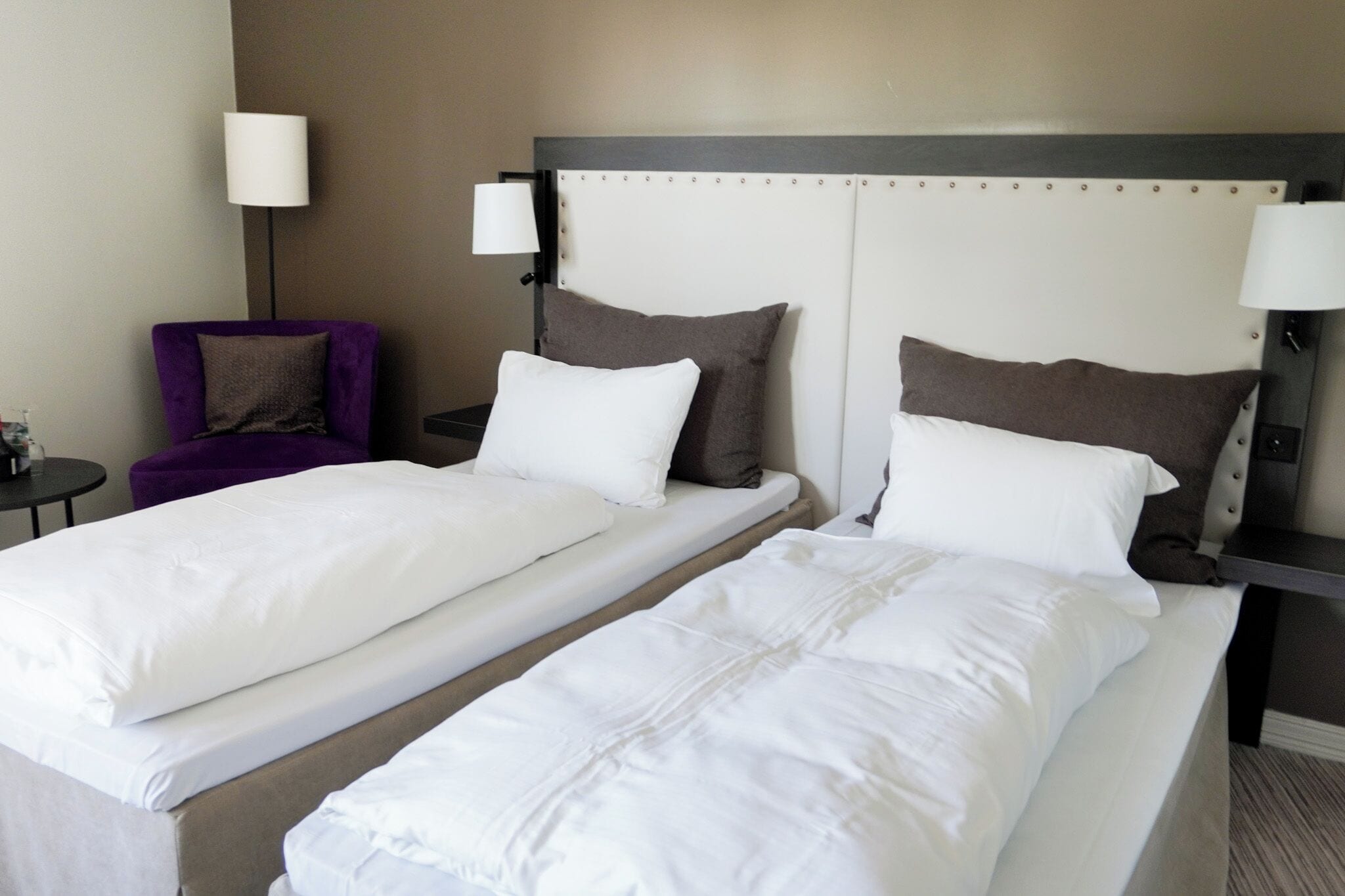 A hotel room with two single beds, a purple armchair, a small table, and a lamp.