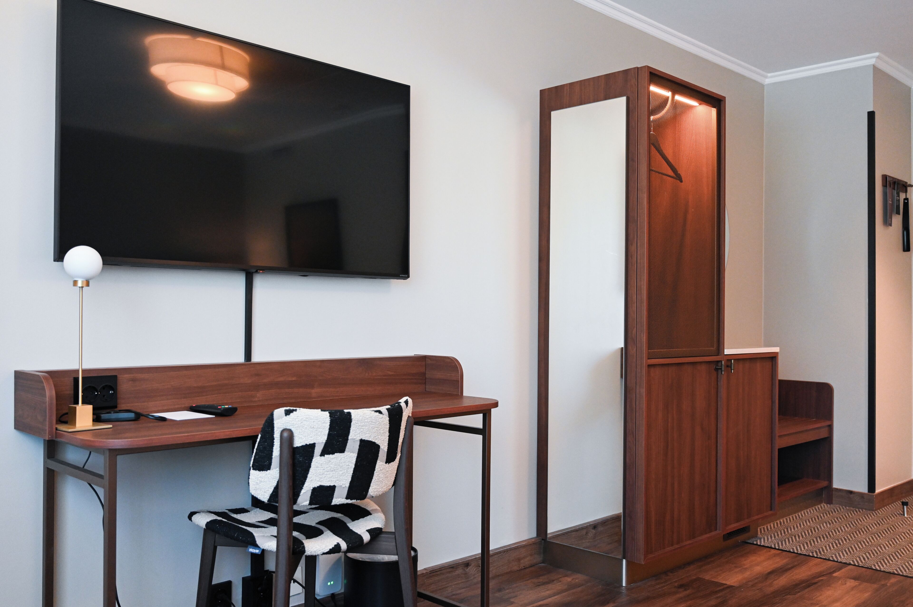 A modern room with a flat-screen TV mounted on the wall, a wooden desk with a chair, a coat closet, and a small seating area with a patterned rug.