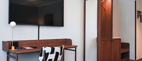 A modern room with a flat-screen TV mounted on the wall, a wooden desk with a chair, a coat closet, and a small seating area with a patterned rug.