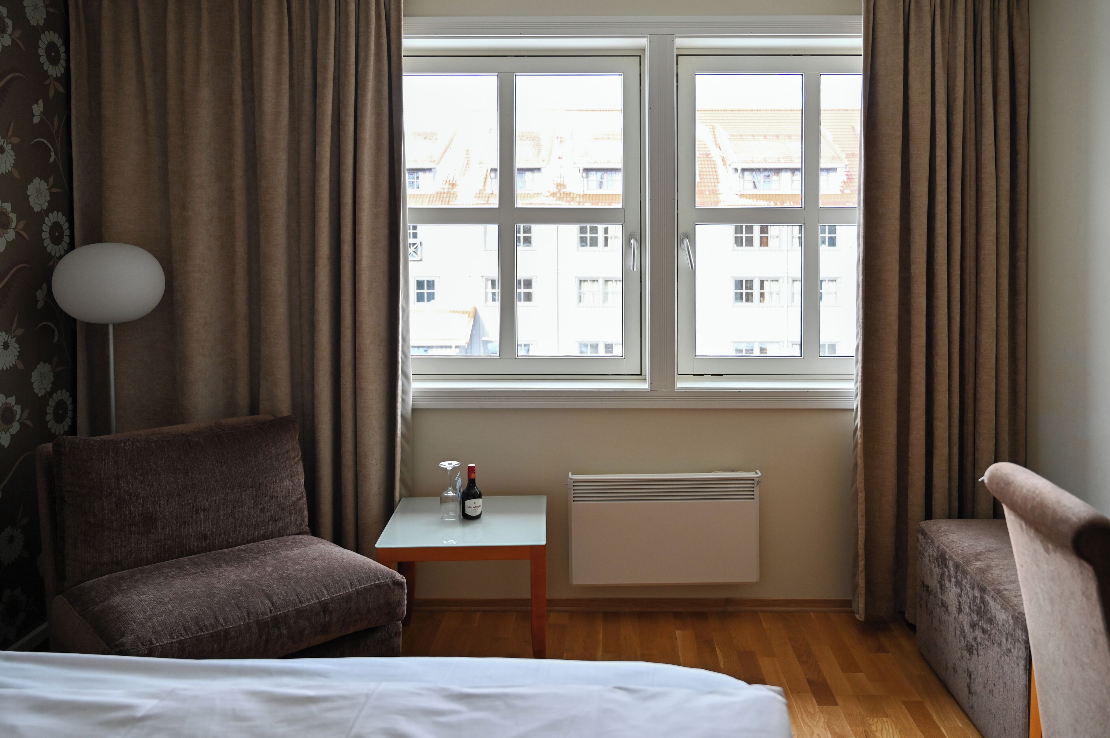 A hotel room with a bed, two armchairs, a small table with a wine bottle and glass, a radiator, and a window with curtains.