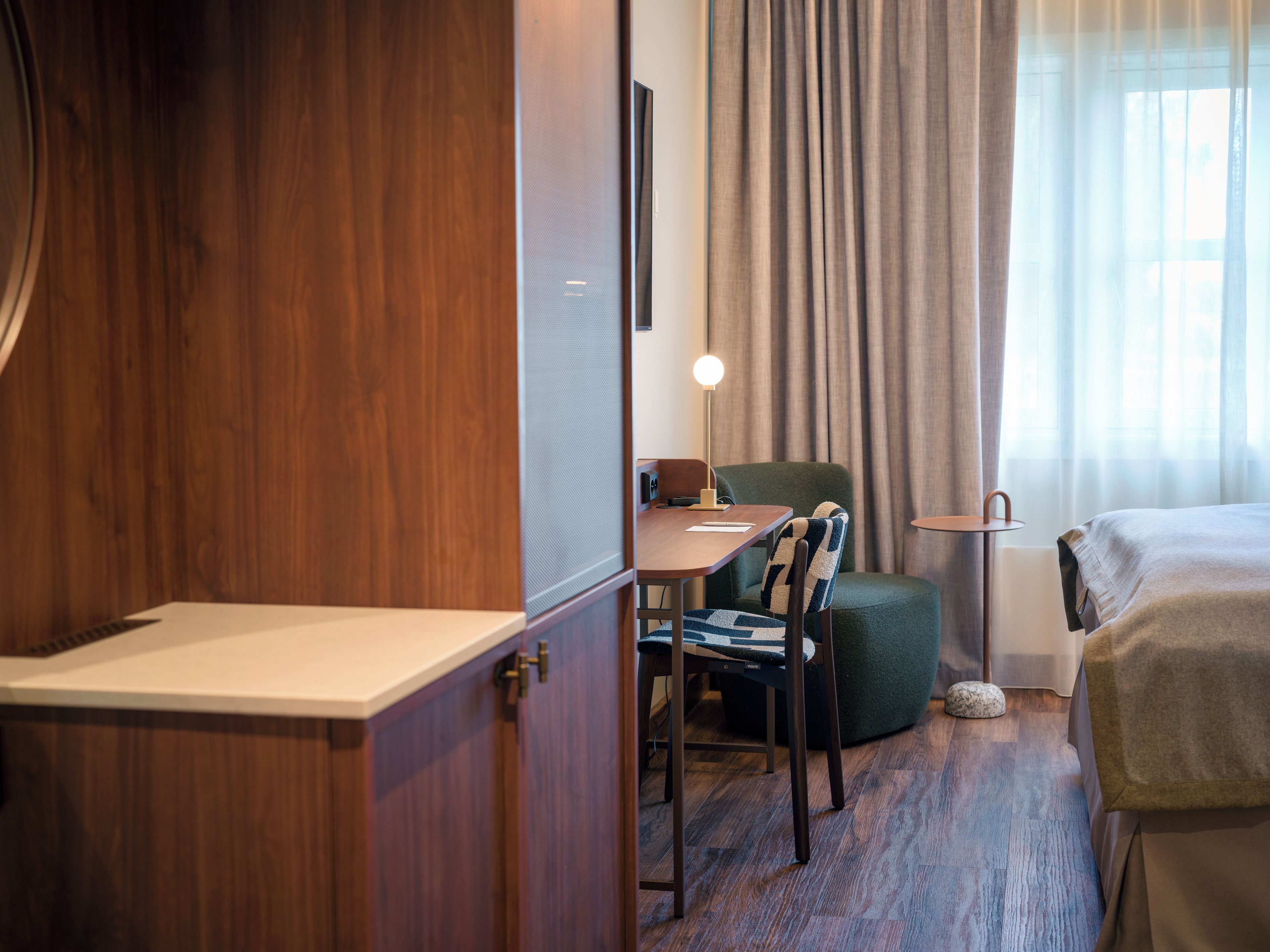 A hotel room with a wooden wardrobe, a desk with a lamp, a green armchair, a bed, and a window with curtains.