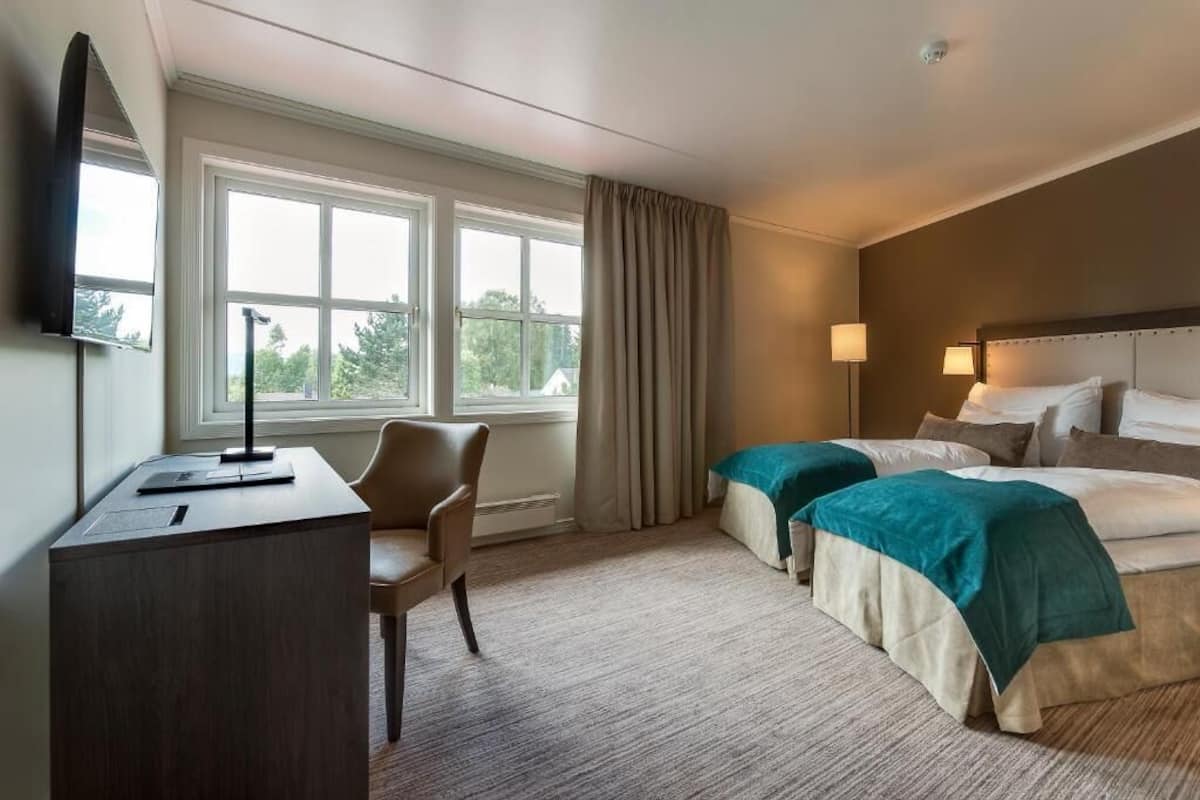 Una habitación de hotel con escritorio, dos camas, una silla y un ventanal grande con cortinas.