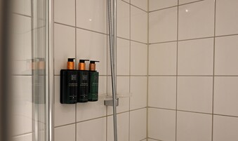 Baño con cabina de ducha de vidrio, paredes de azulejos blancos y un juego de artículos de aseo en un estante.