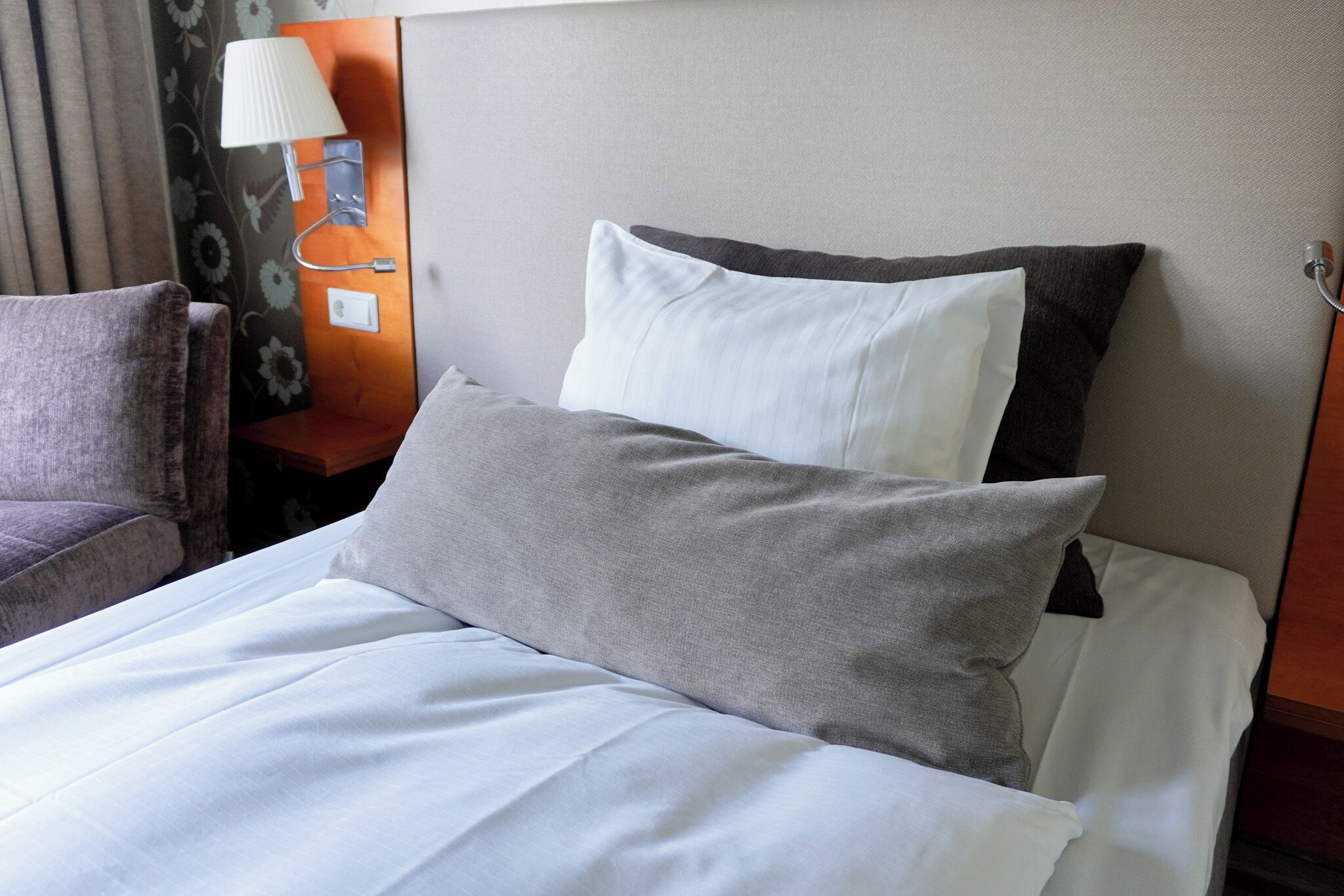 A neatly made bed with white and gray linens, a nightstand with a lamp, and a floral-patterned curtain.