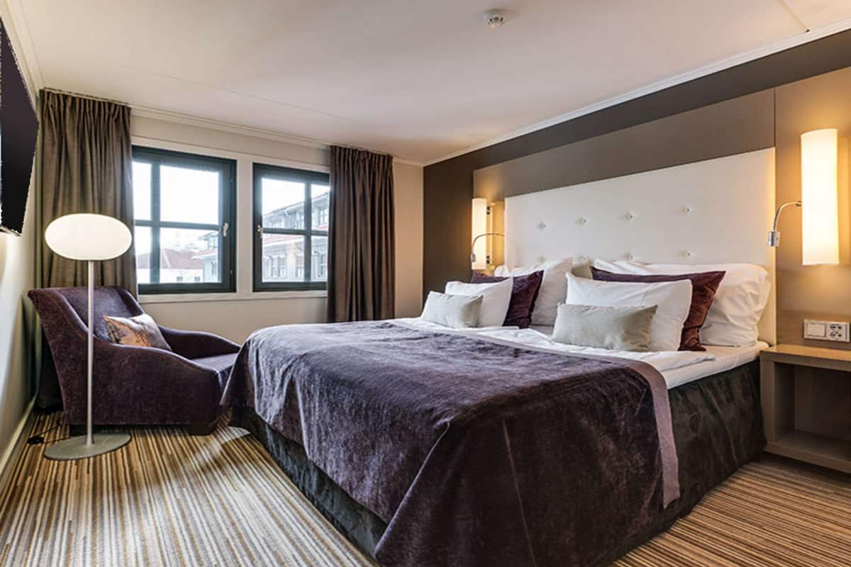 Habitación de hotel con una cama grande, una silla, una lámpara y vistas a edificios a través de la ventana.