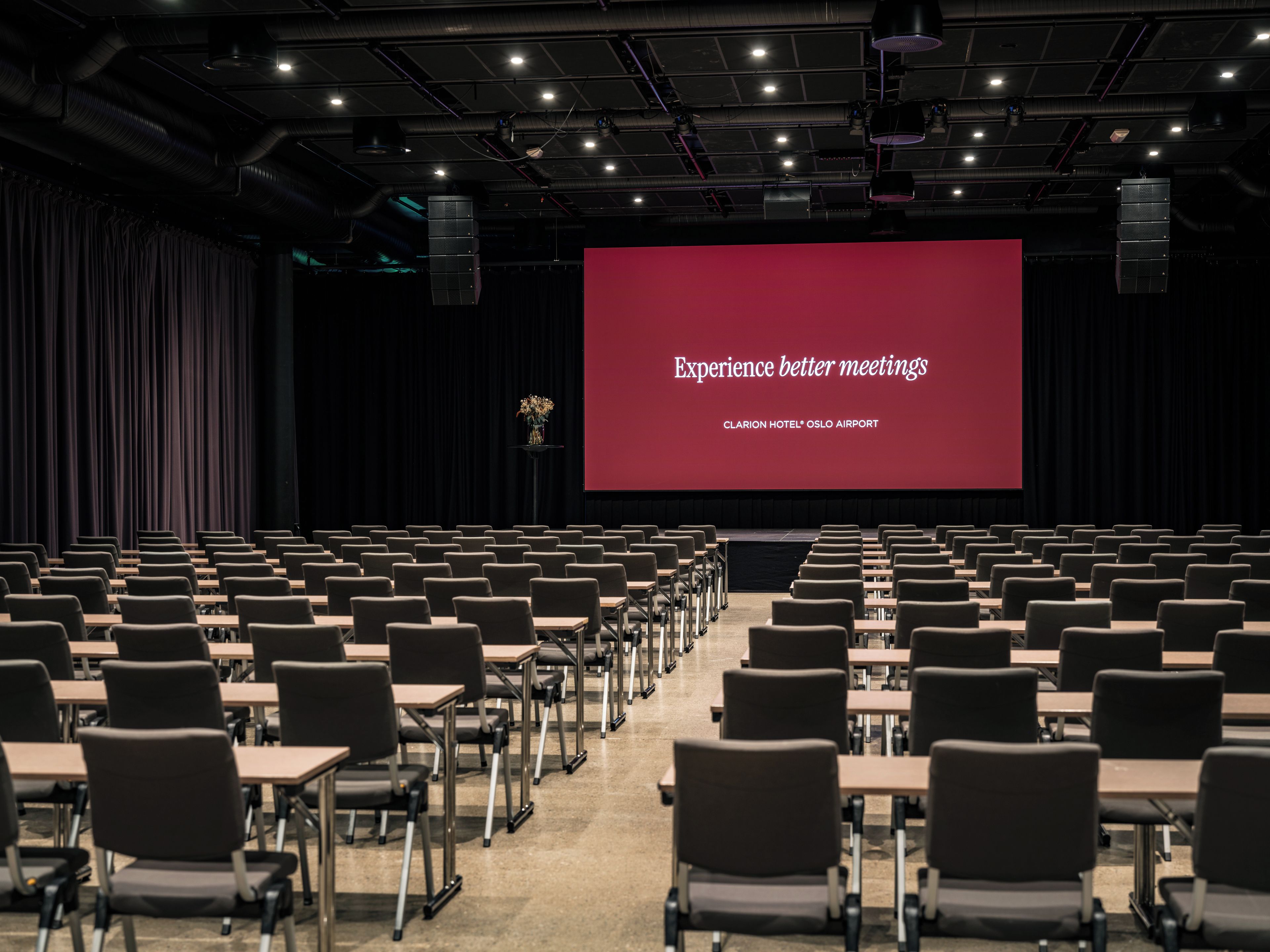 A conference room with rows of chairs and a large screen displaying 'Experience better meetings' at Clarion Hotel Oslo Airport.