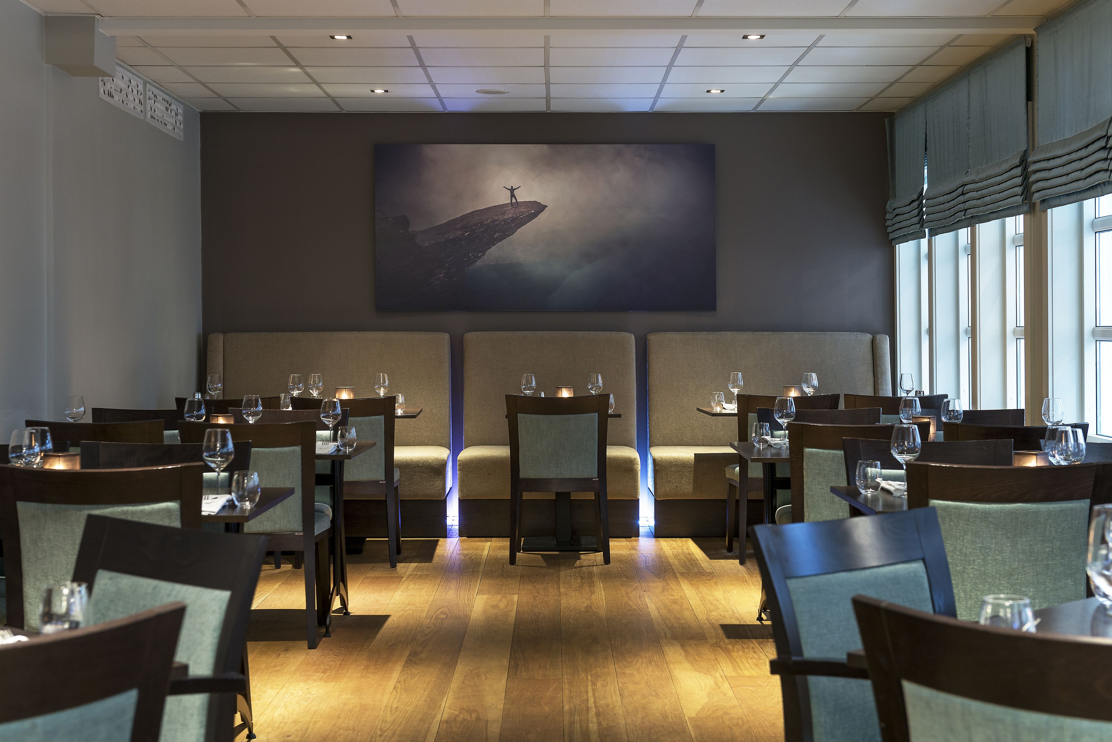 A restaurant with wooden tables and chairs, a large painting on the wall, and a view of the city through large windows.