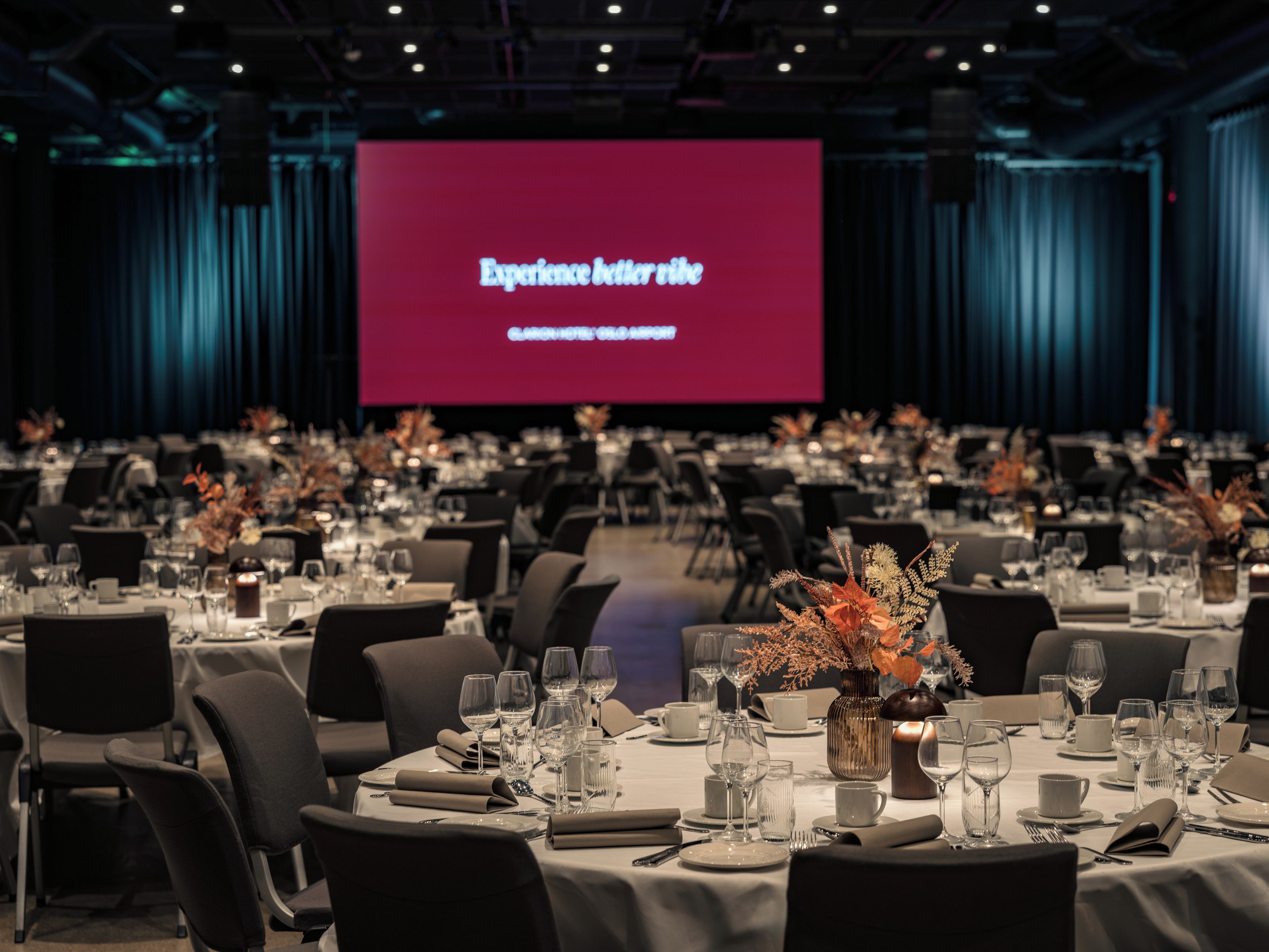 A conference room with tables set for a formal event, a large screen displaying text, and a stage area.
