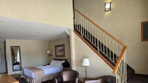 A hotel room with a bed, two chairs, a desk, a lamp, a painting, and a staircase. - Lakeview Inns & Suites - Hinton (Hinton)