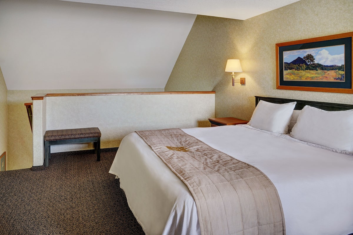 Habitación de hotel con una cama grande, un banco, una mesita de noche, una lámpara y un cuadro en la pared.