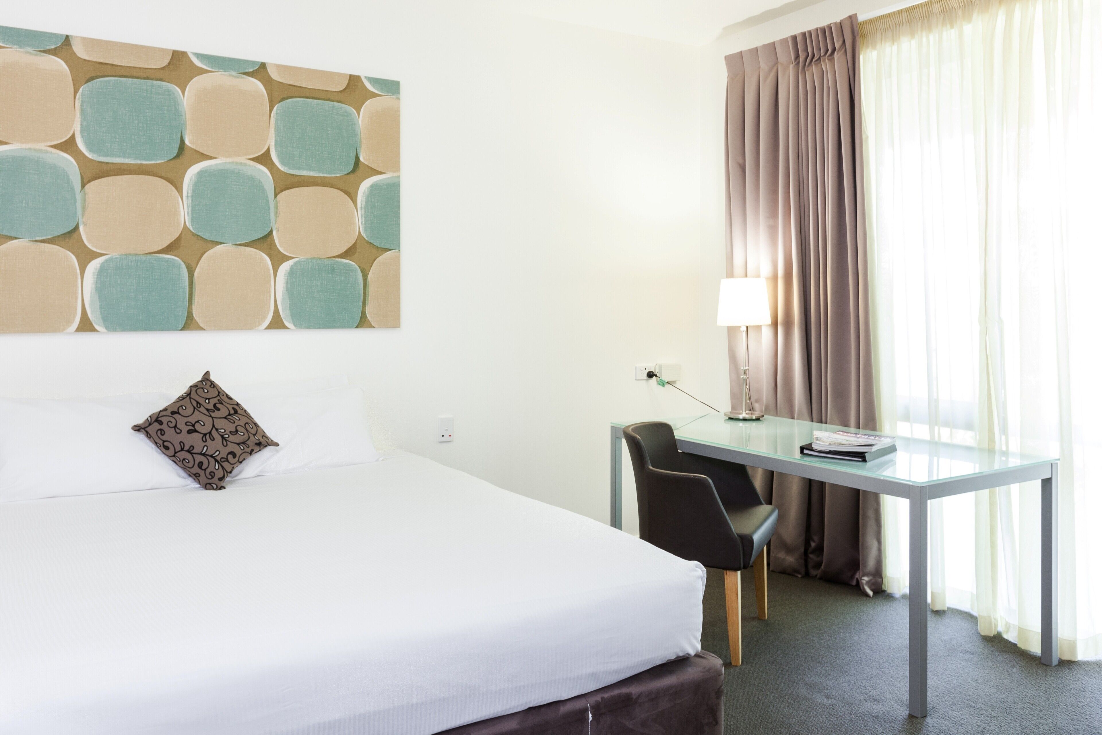 A hotel room with a bed, a desk, a chair, a lamp, and a window with curtains.