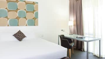 Et hotelværelse med seng, skrivebord, stol, lampe og et vindue med gardiner.