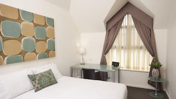 A hotel room with a bed, a desk, a chair, a lamp, a window with curtains, and a small table with a plant.