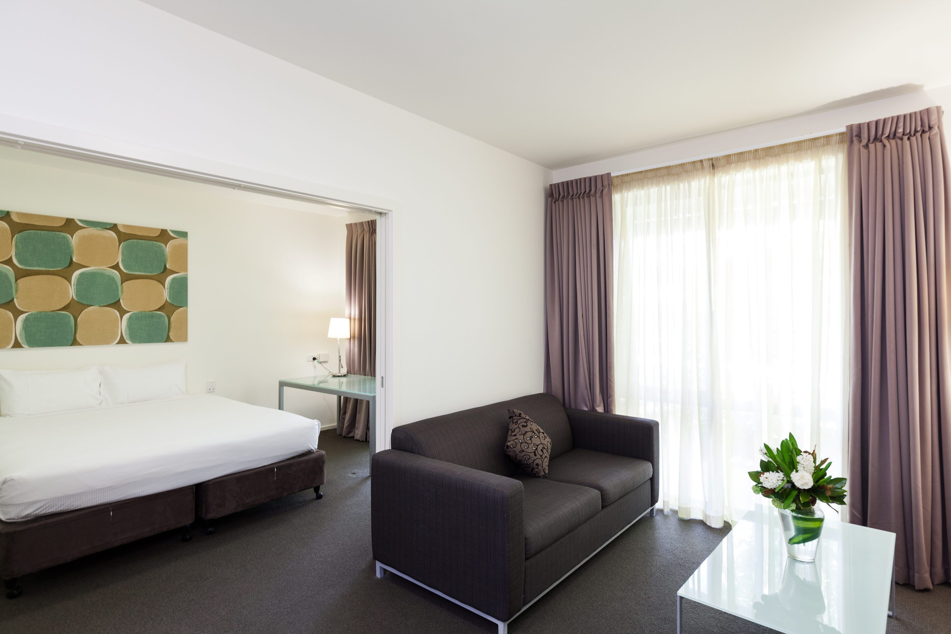 A hotel room with a bed, a sofa, a small table, a lamp, and a window with curtains.