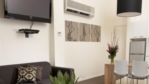 Una sala moderna con televisión de pantalla plana, una mesa de centro de vidrio, un sofá con cojines estampados, un comedor con mesa y sillas, y un aire acondicionado instalado en la pared.
