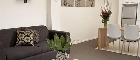 A modern living room with a flat-screen TV, a glass coffee table, a sofa with patterned cushions, a dining area with a table and chairs, and a wall-mounted air conditioner.