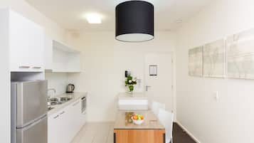 A modern kitchen with white cabinets, a stainless steel refrigerator, a sink, and a wooden dining table with a bowl of fruit.