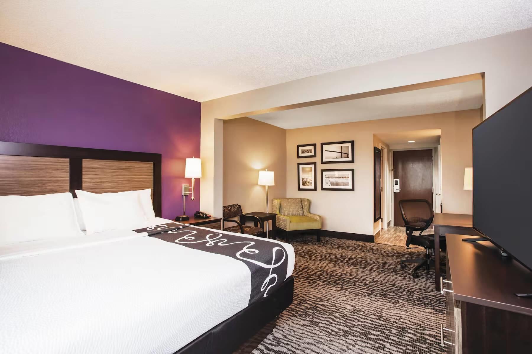 A hotel room with a bed, a desk, a chair, a lamp, and framed pictures on the wall.