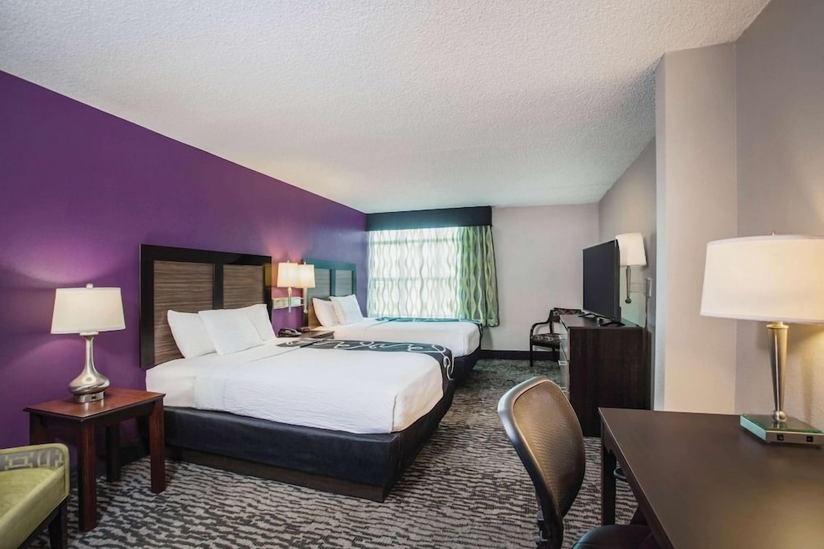 A hotel room with two beds, a desk, a chair, a TV, and a window with curtains.