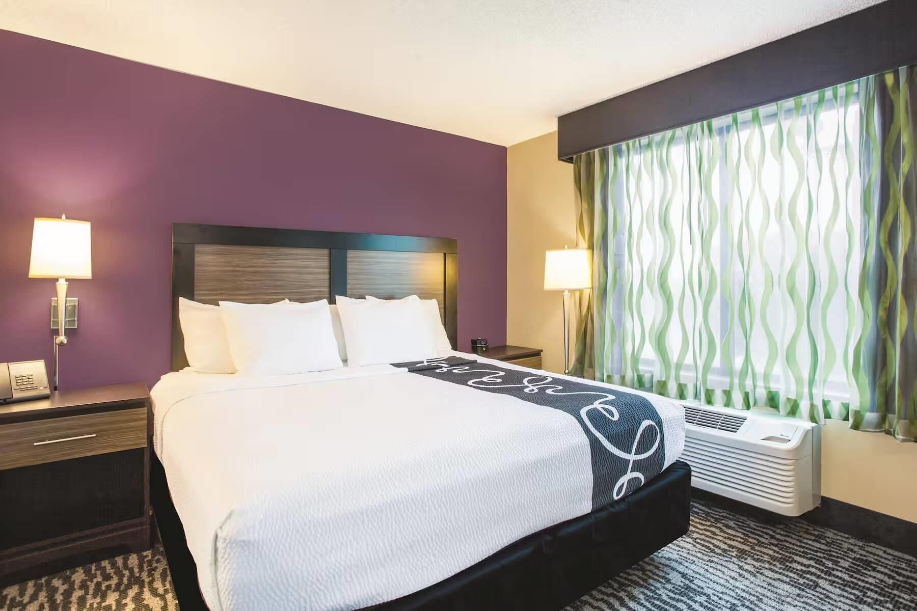 A hotel room with a bed, two bedside tables, a nightstand, a window with curtains, and an air conditioning unit.