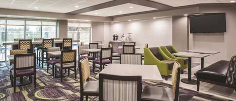 A hotel dining area with multiple tables and chairs, a flat-screen TV mounted on the wall, and a large window providing natural light.