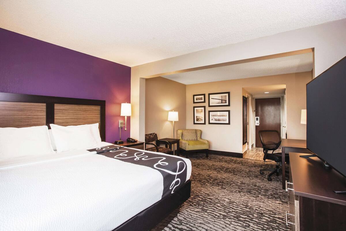 A hotel room with a bed, a desk, a chair, a TV, and a door leading to another room.