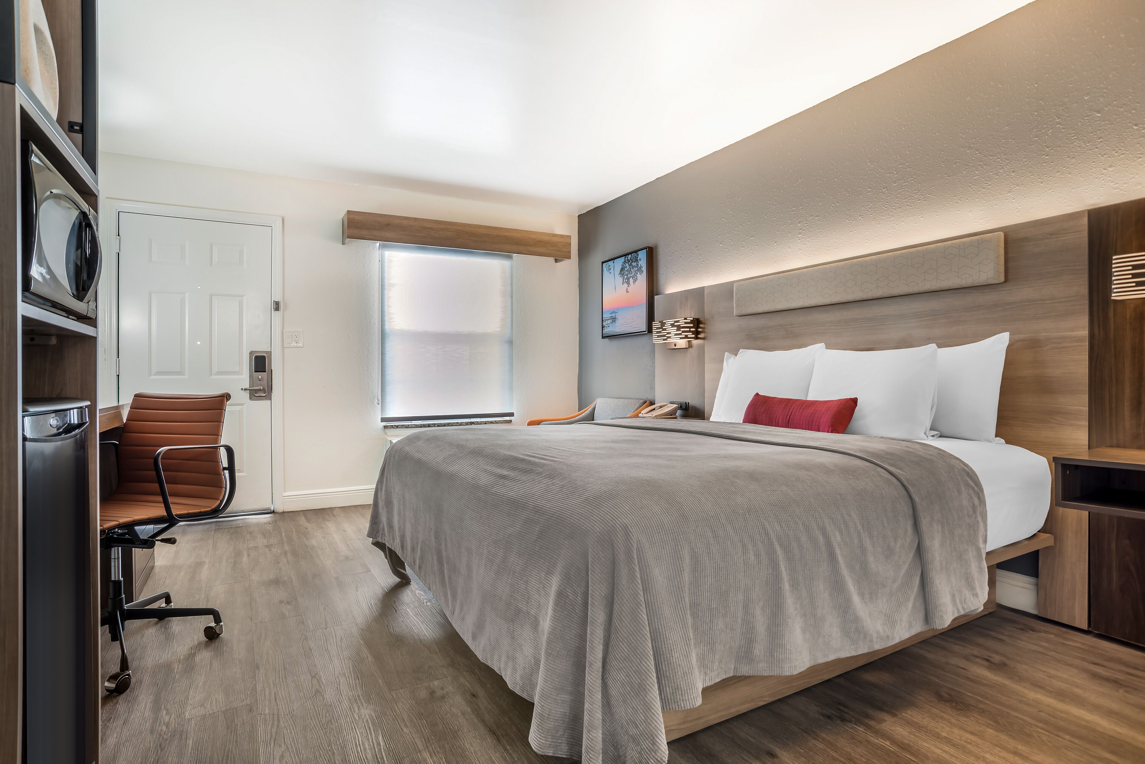 A modern hotel room with a large bed, a desk with a chair, a microwave, and a window with blinds.