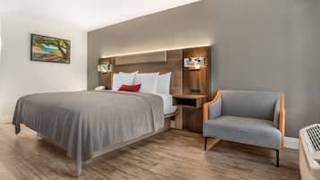 A modern hotel room with a large bed, a grey armchair, a nightstand with a lamp, and a painting on the wall.