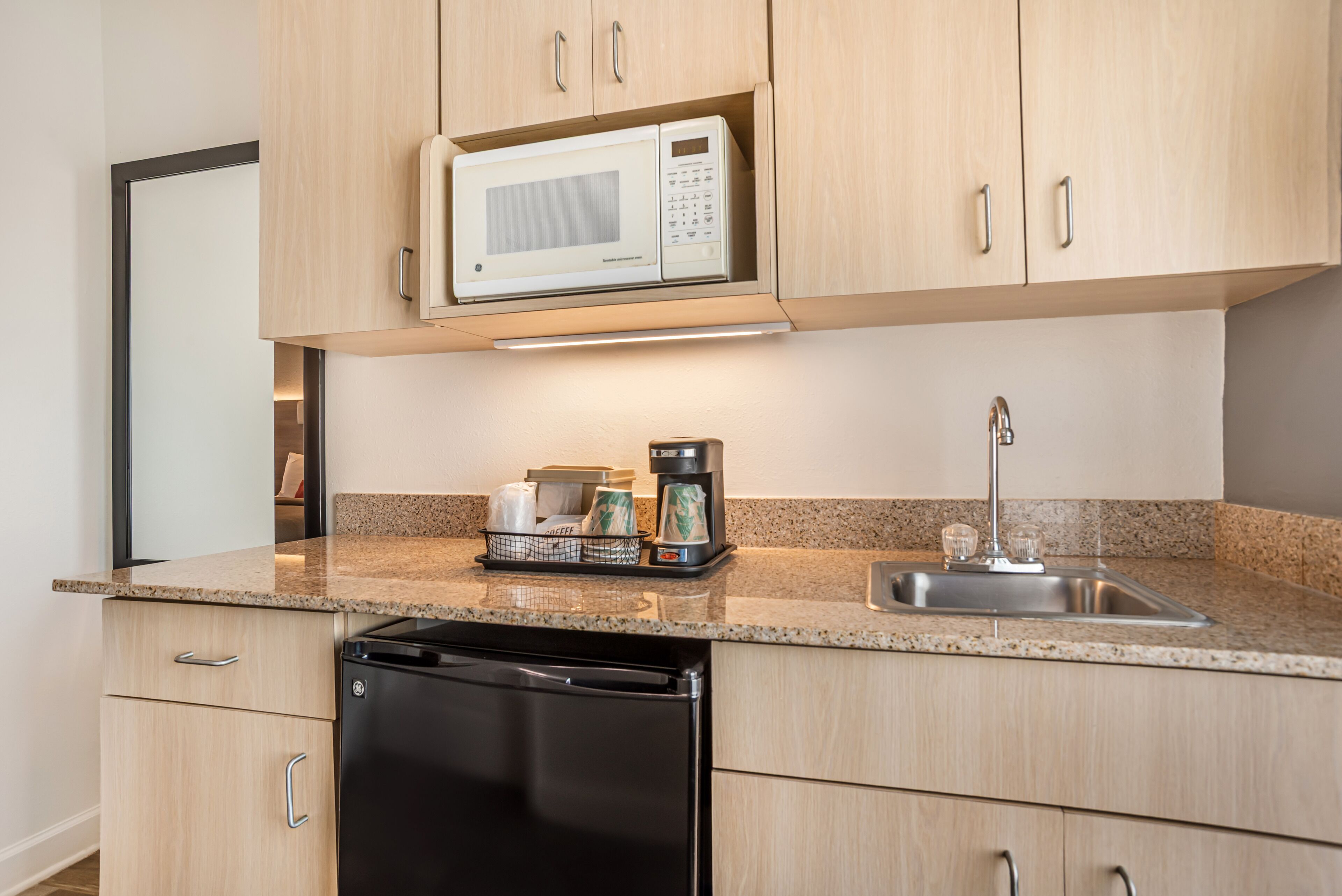 A modern kitchen with light wood cabinets, a built-in microwave, a coffee maker, and a dishwasher.