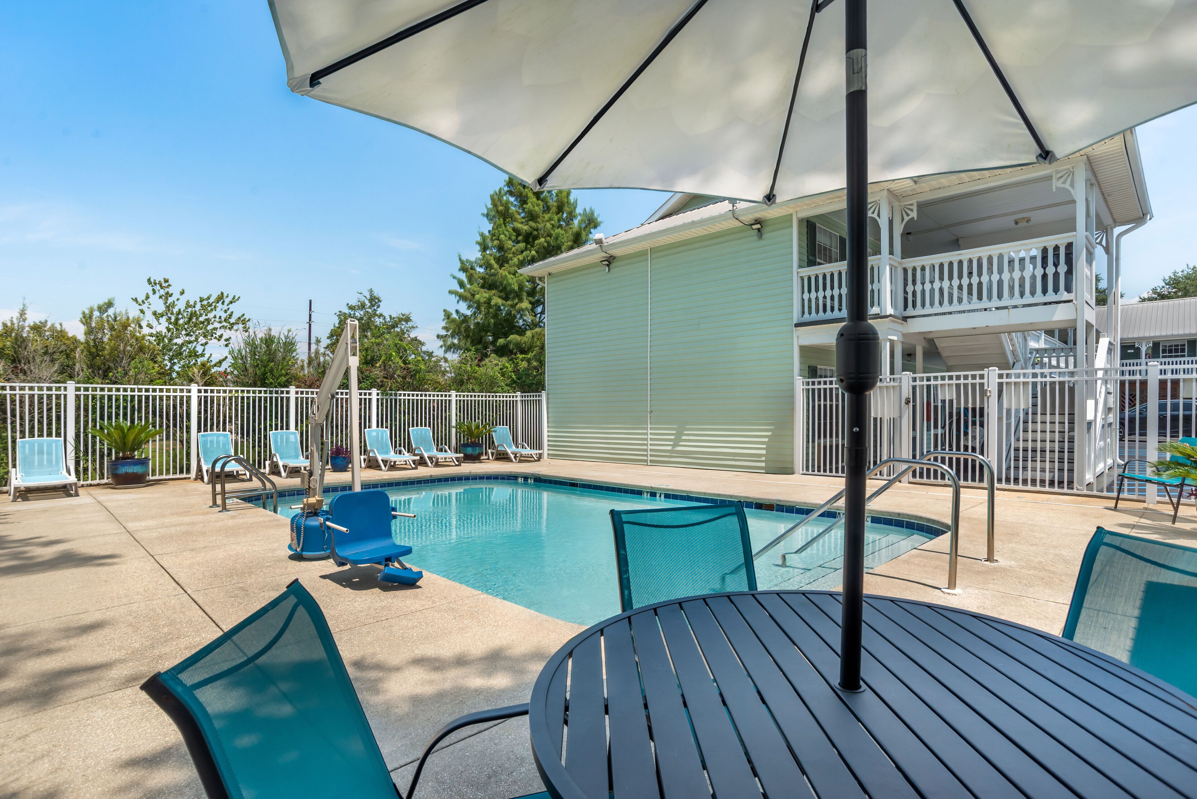 A swimming pool with lounge chairs and a table, surrounded by a white fence and a building with a balcony.