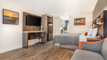 A modern hotel room with a wooden desk, a flat-screen TV, a bed with grey and orange bedding, and artwork on the walls.