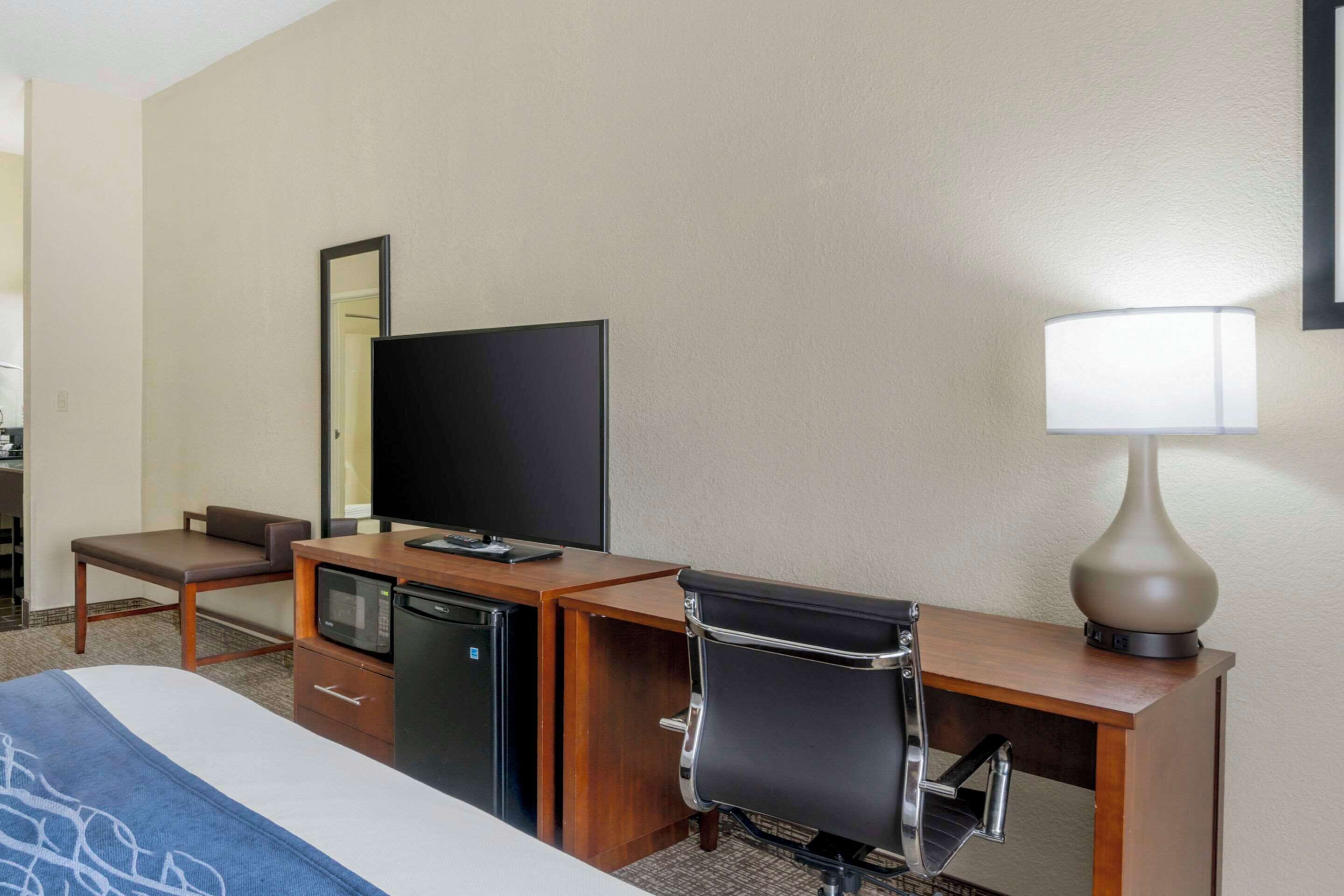 A hotel room with a bed, a desk with a computer, a microwave, a chair, a TV, and a lamp.