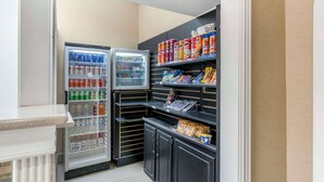 A compact kitchenette with two refrigerators stocked with beverages and snacks, and shelves with packaged goods. - Comfort Inn & Suites La Grange - Louisville Area (La Grange)
