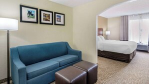 A hotel room with a blue sofa, a brown ottoman, a bed, and framed pictures on the wall. - Comfort Inn & Suites La Grange - Louisville Area (La Grange)