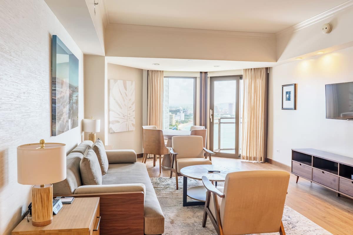 A modern hotel room with a sofa, armchairs, a coffee table, and a TV.