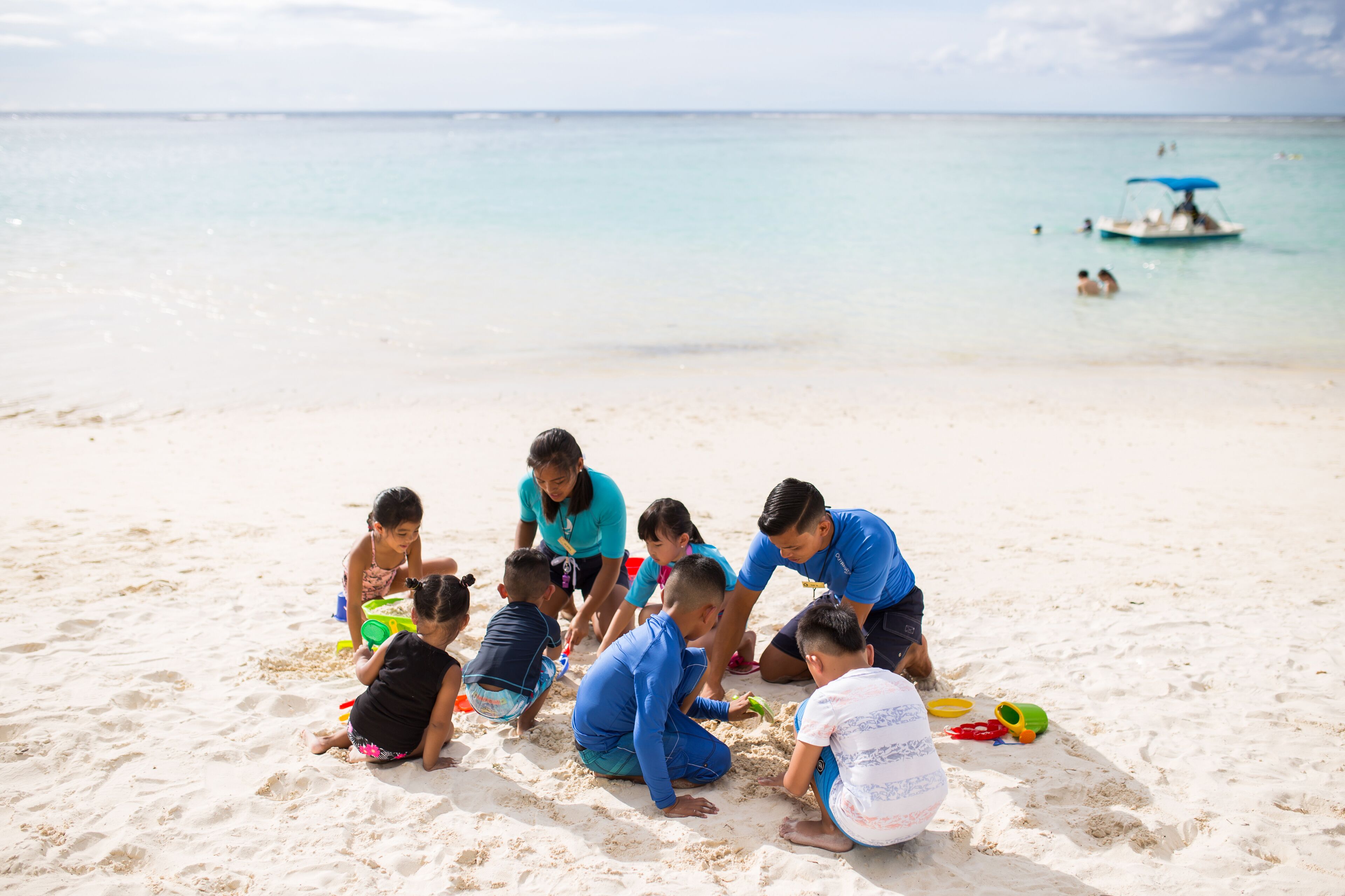 A group of children and adults on a sandy beach with a boat in the background.