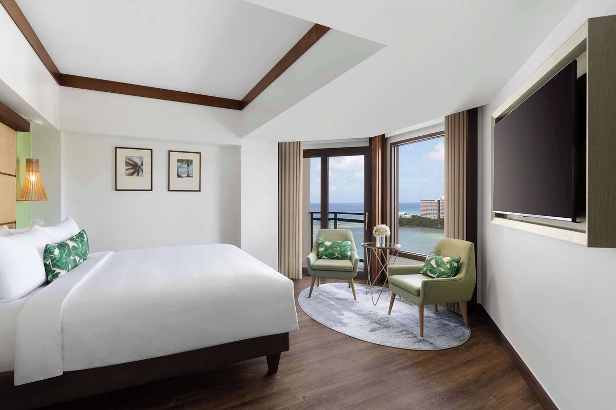 A hotel room with a large bed, two chairs, a TV, and a view of the sea.